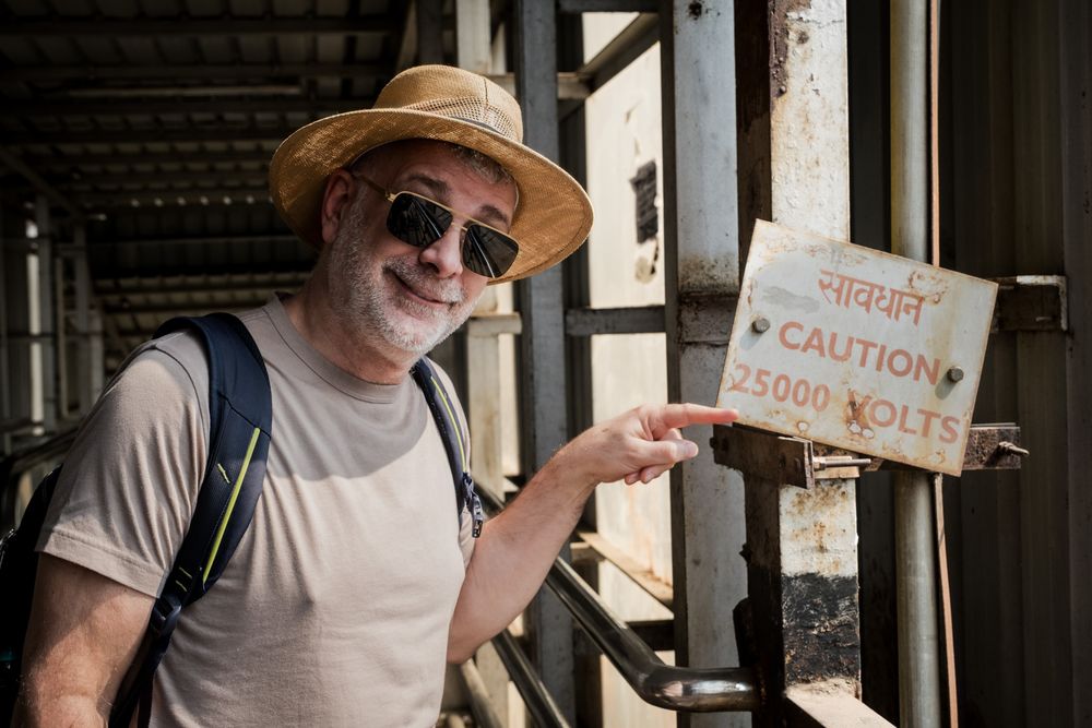 Tourist pointing at a caution 25000 volts warning sign on a rustic metal structure in India.