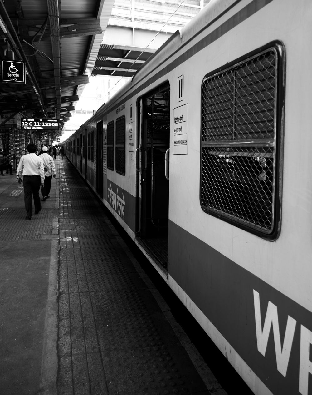 Black and white image of passengers walking alongside a train on an Indian railway platform.