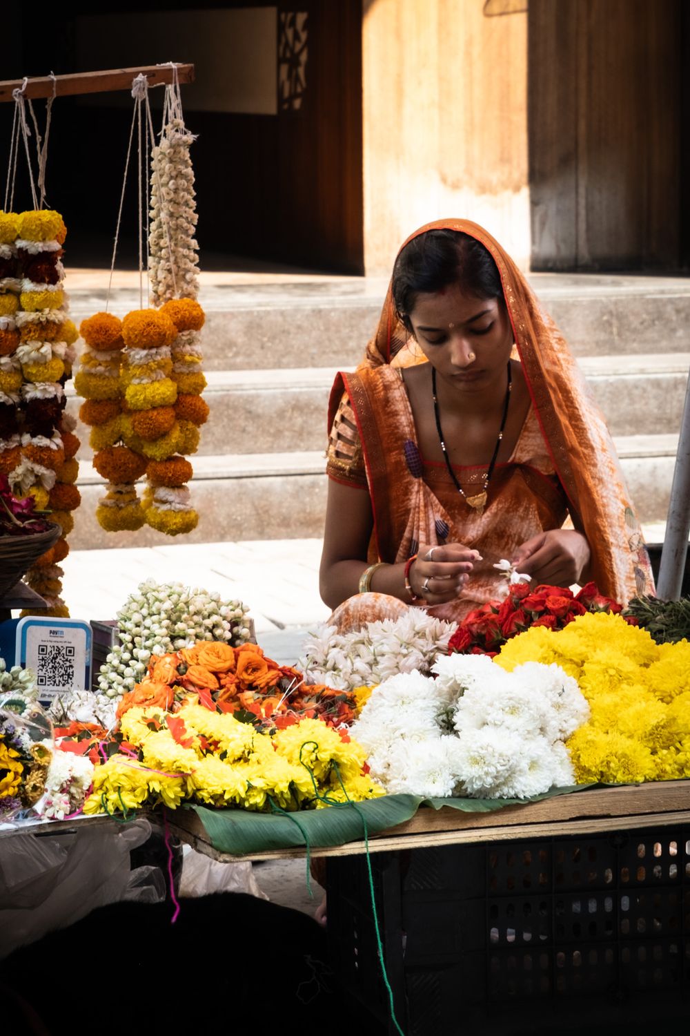 Indian woman in orange sari arranging colorful flower garlands at a street market stall.