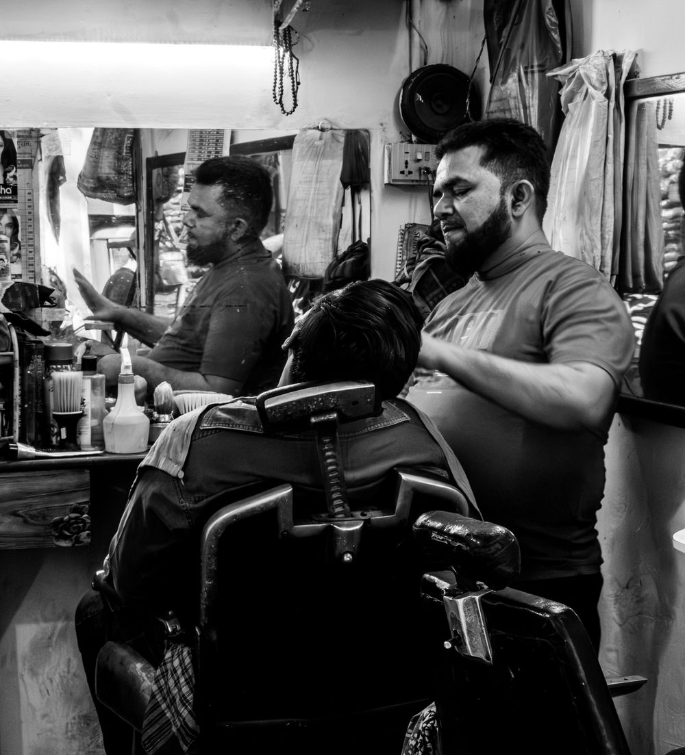 Black and white photo of barber cutting hair in a traditional Indian barbershop.
