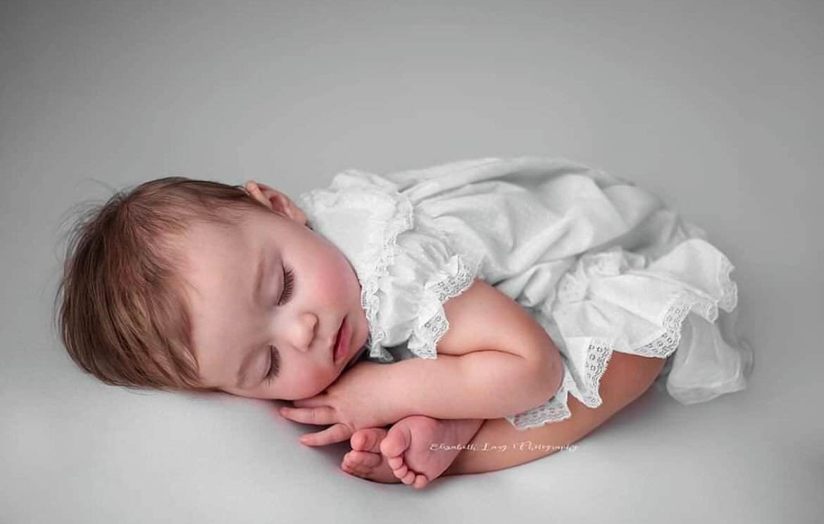 A sleeping baby in a white lace dress curls up peacefully on a soft grey background.