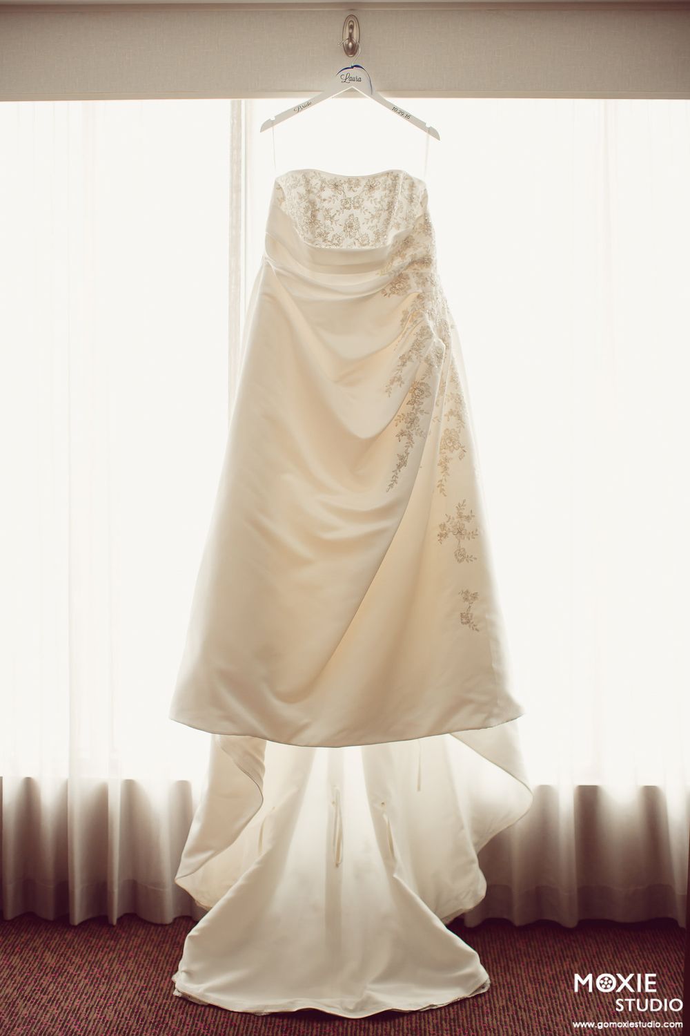 A white wedding dress with delicate embellishments hangs elegantly against a window, creating a soft silhouette.