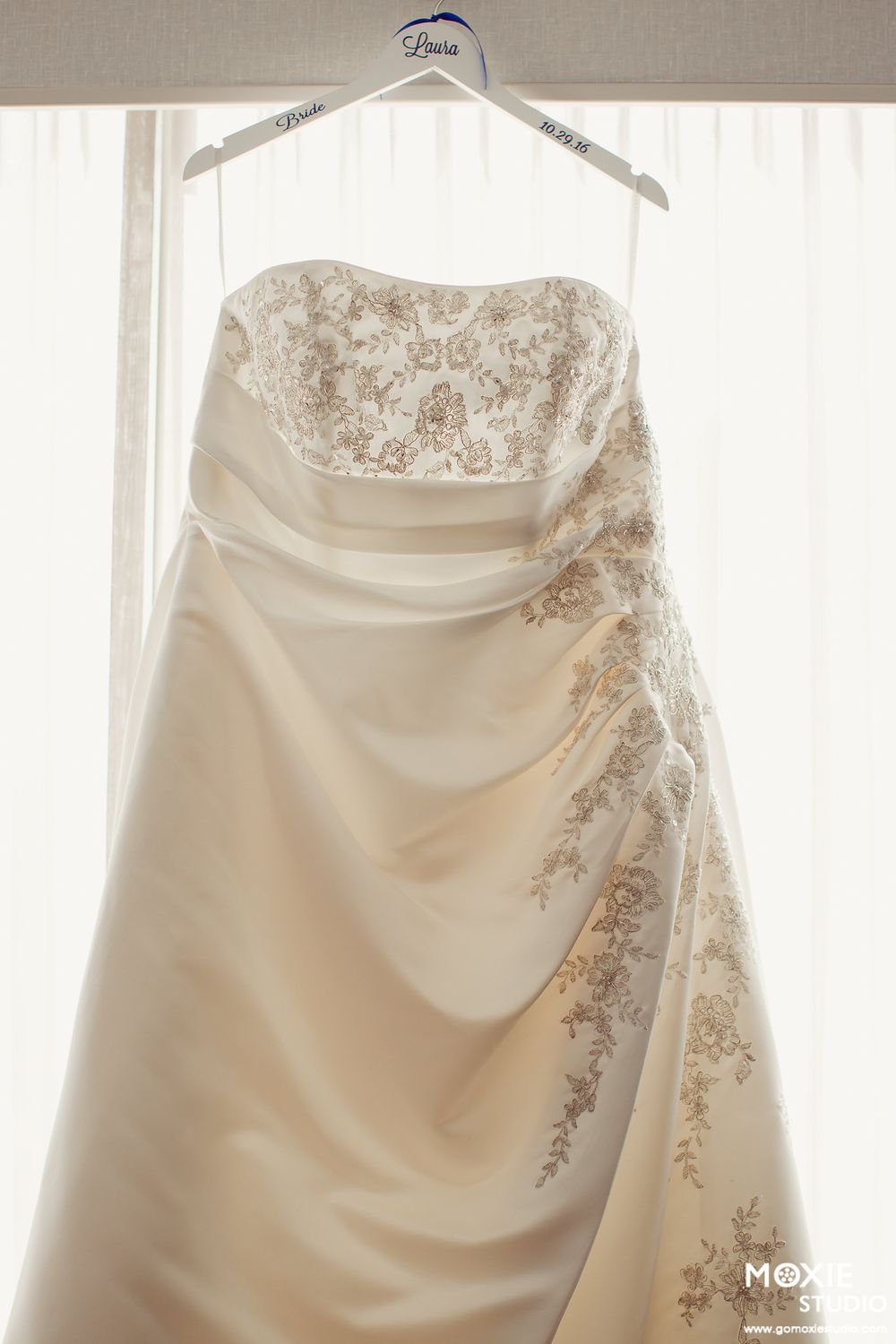 Close-up view of a white wedding dress with delicate beaded embroidery details hanging against a light window backdrop.