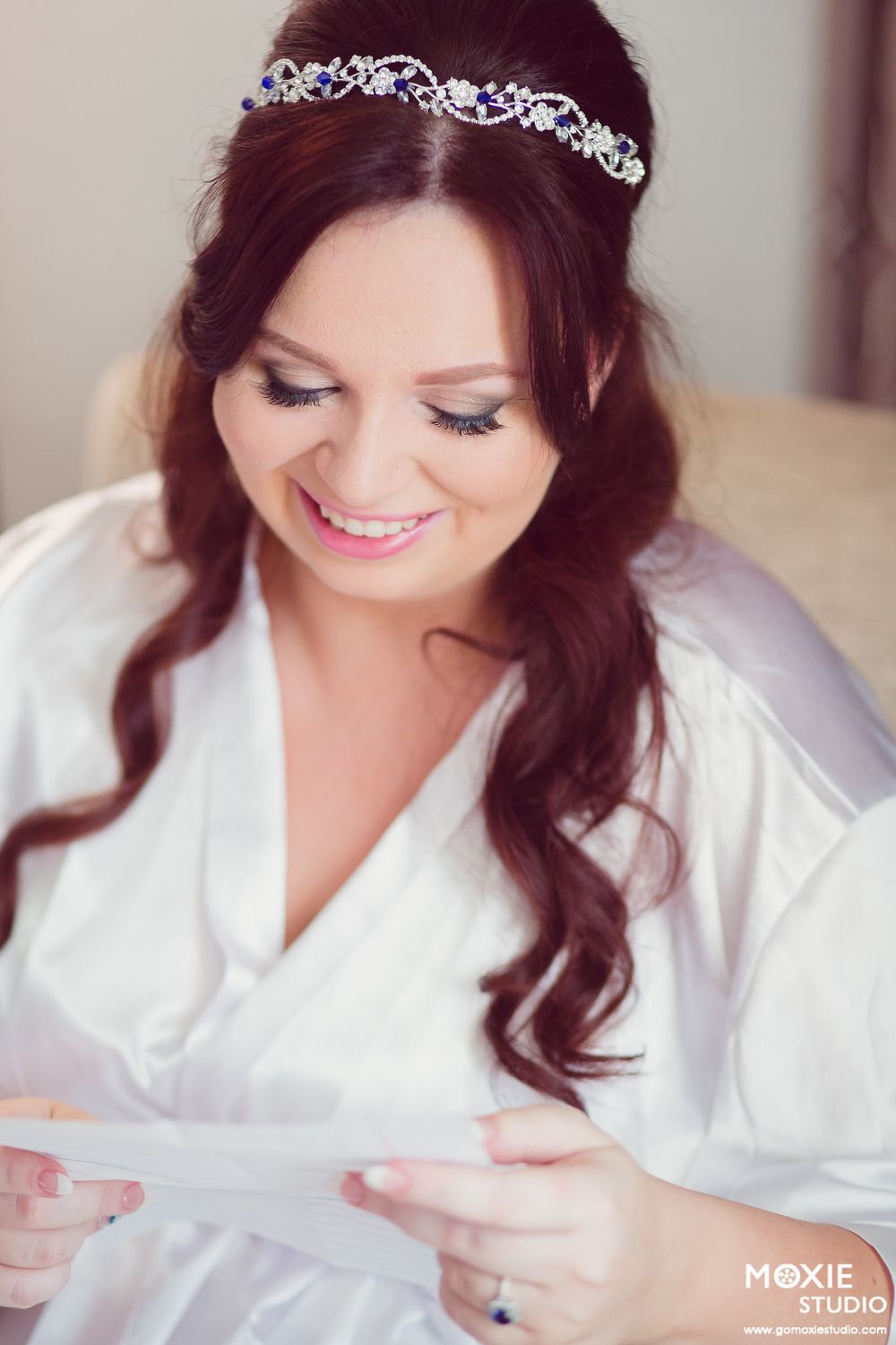 A bride wearing a delicate crystal tiara and white robe smiles while looking down during wedding preparations.