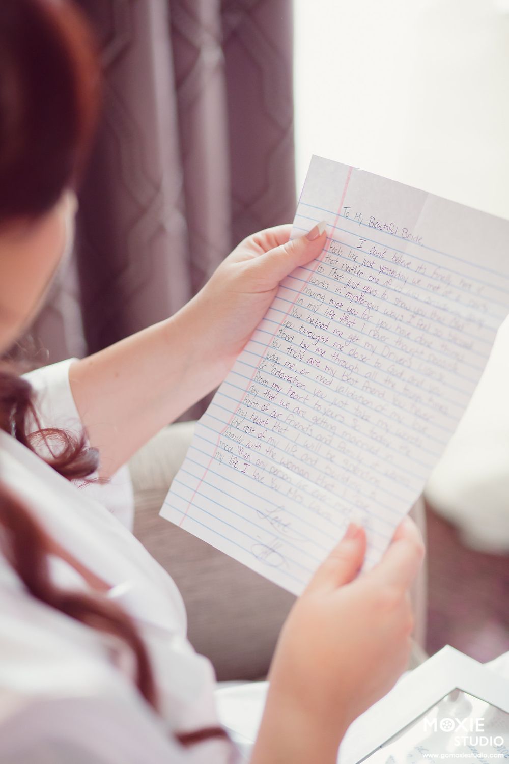 Hands holding a handwritten letter on lined paper in a softly lit room with curtains in the background.