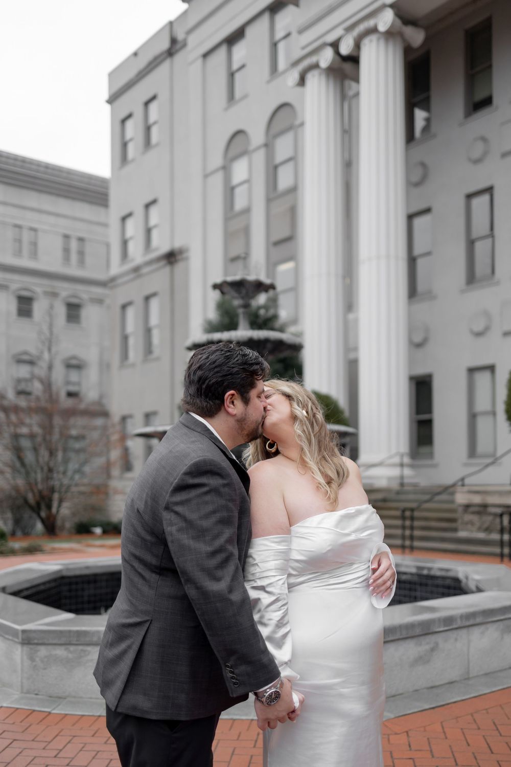 A romantic wedding couple shares a tender kiss on a brick patio in front of a stately white columned building.