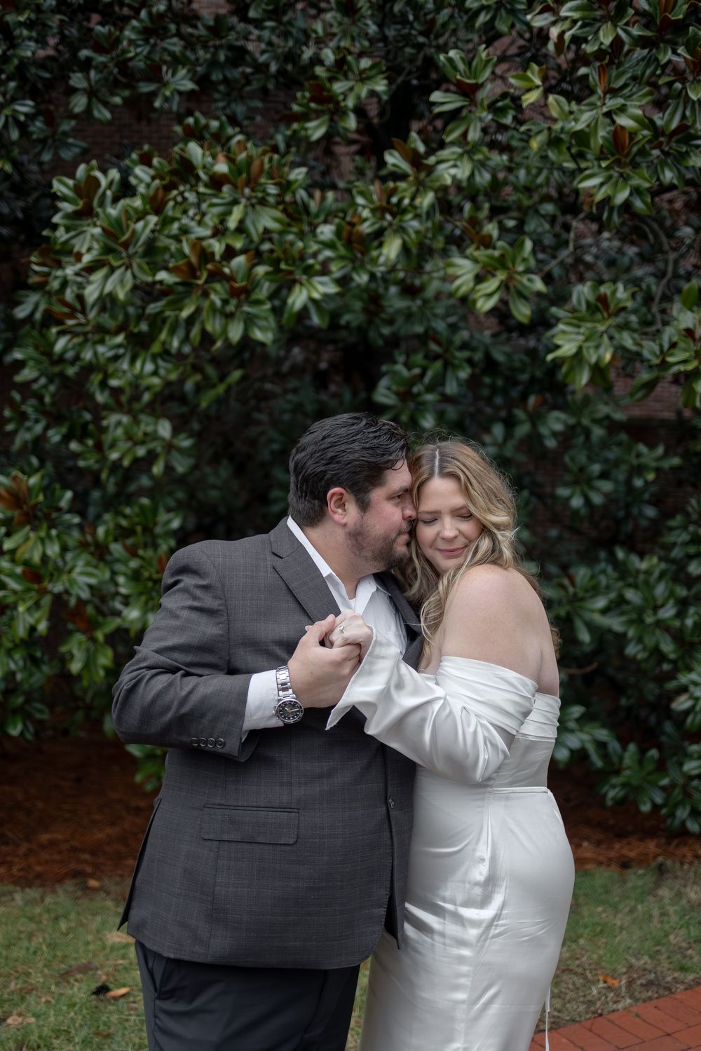 A couple in formal wedding attire share an intimate moment together against a backdrop of lush green foliage.