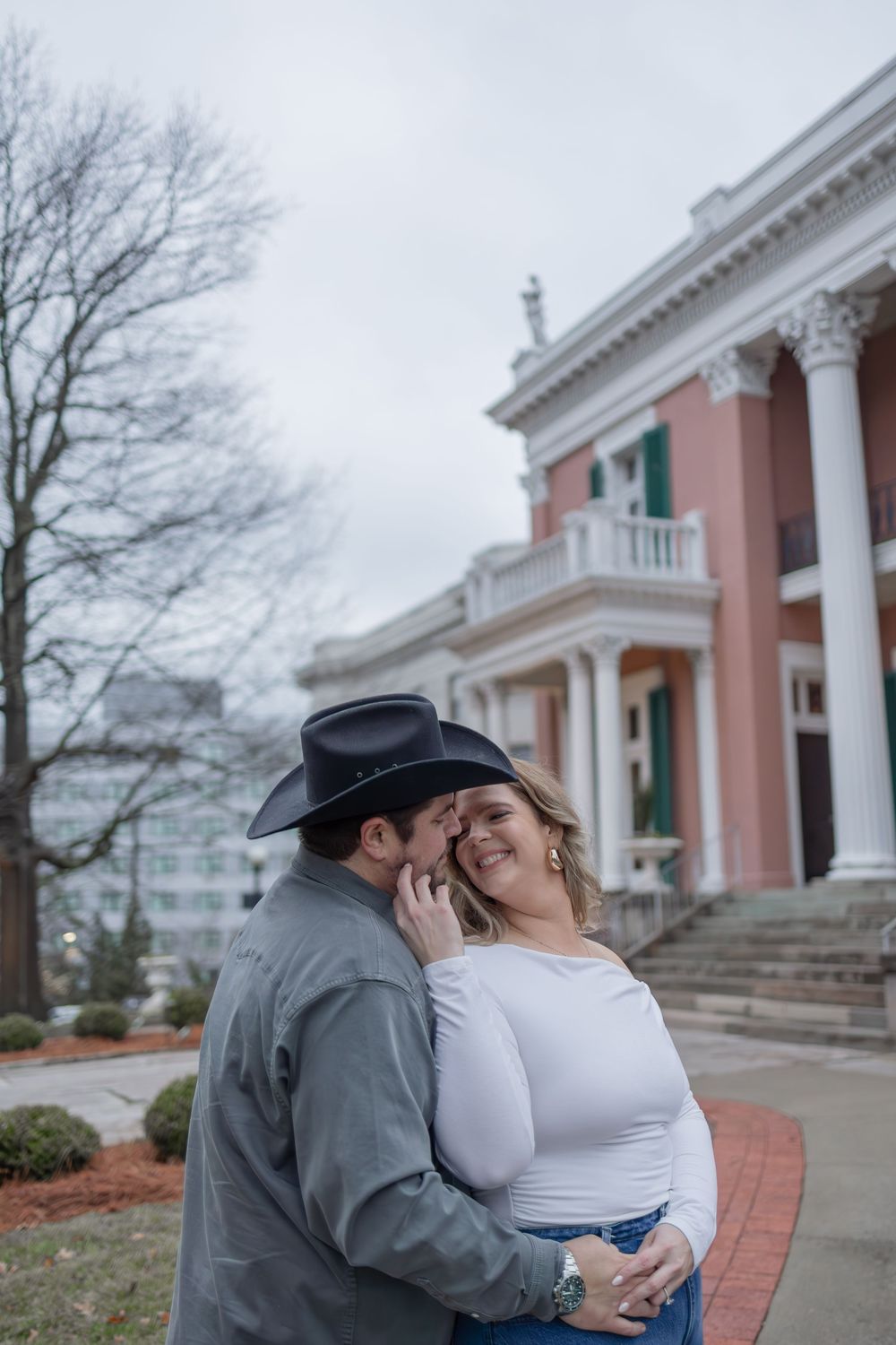 A couple shares an intimate moment outside a grand pink mansion with white columns on an overcast day.