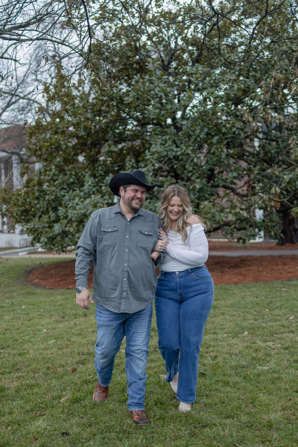 A couple walks together on a grassy lawn in casual attire with a leafy tree behind them on an overcast day.