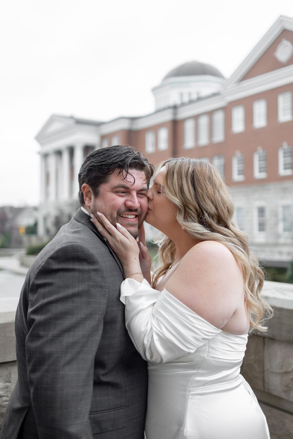 A couple shares a romantic moment on a balcony with brick buildings and white columns in the background.
