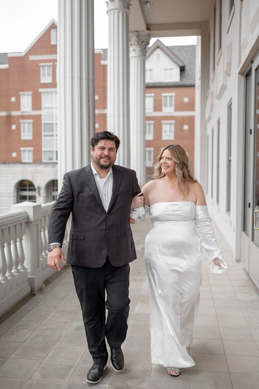 A couple walking together along a covered outdoor walkway with white columns and brick buildings.