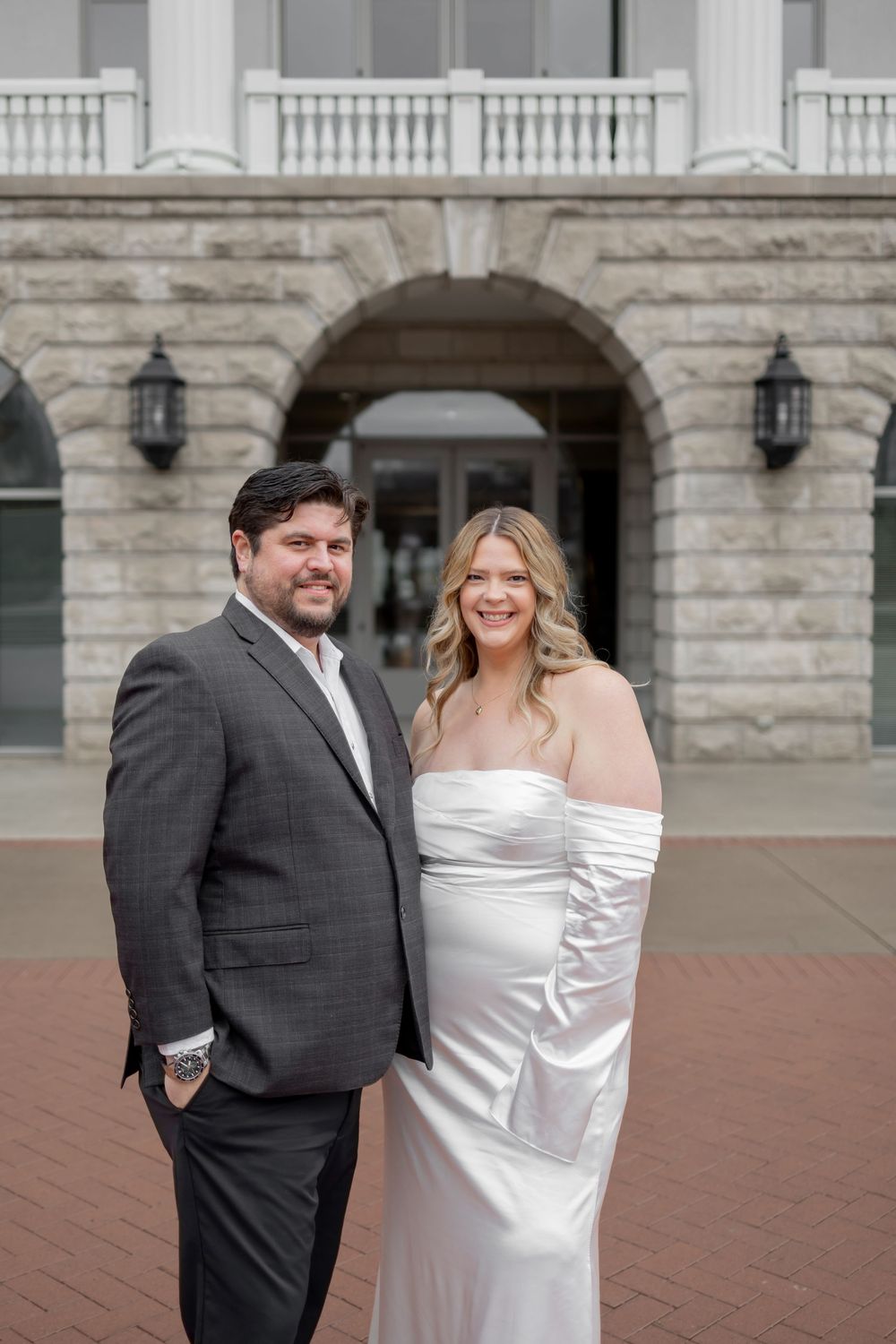 A couple poses together outdoors in formal attire with a white dress and dark suit against a stone building backdrop.