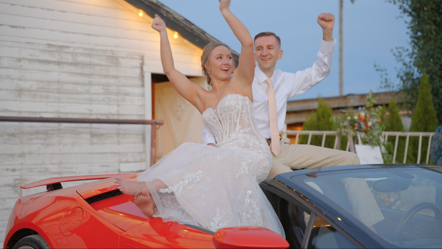 Bride and groom celebrating on top of a red sports car in front of a rustic barn in Des Moines, Iowa.