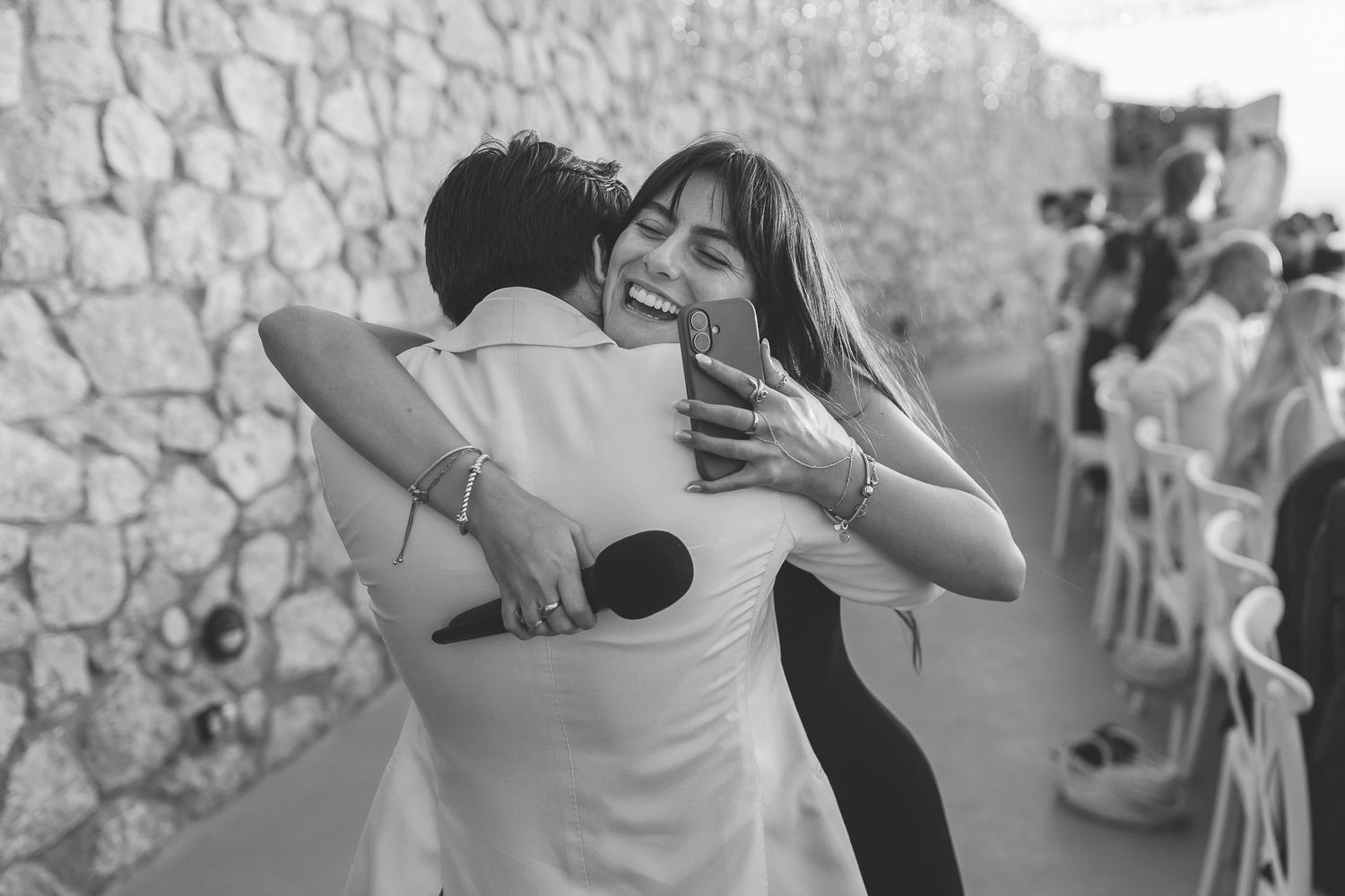 A black and white photo of a Santorini groom and a bridesmaid embracing in front of a stone wall at El Viento.