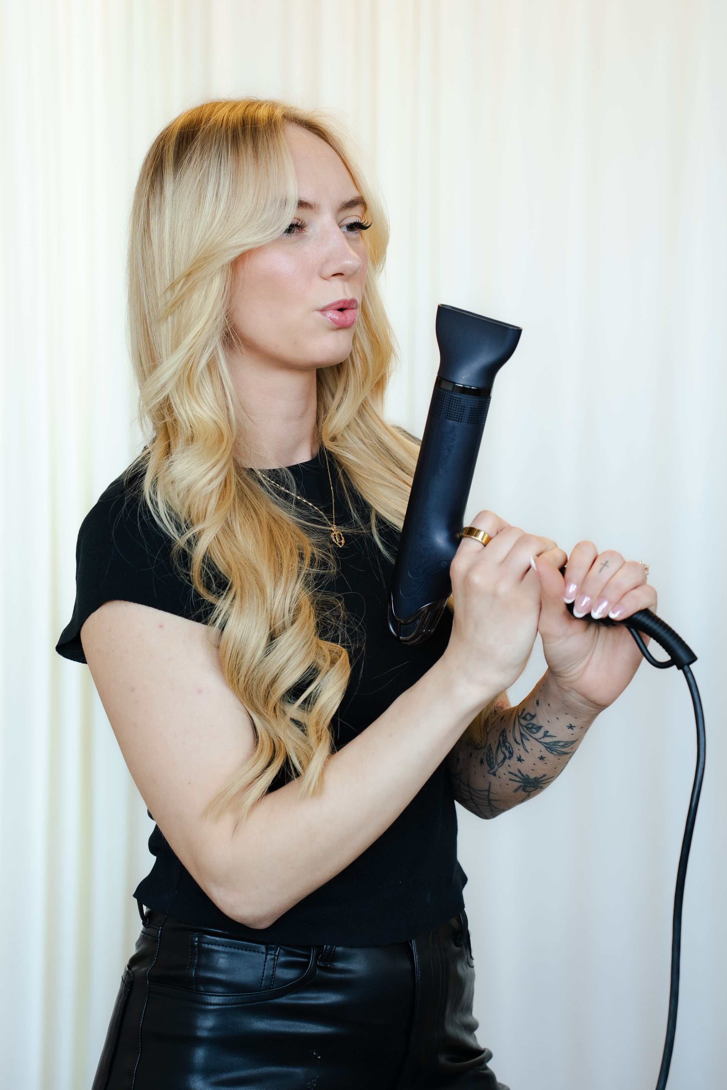 A person with blonde hair poses playfully with a hair dryer against a white background in a black outfit.