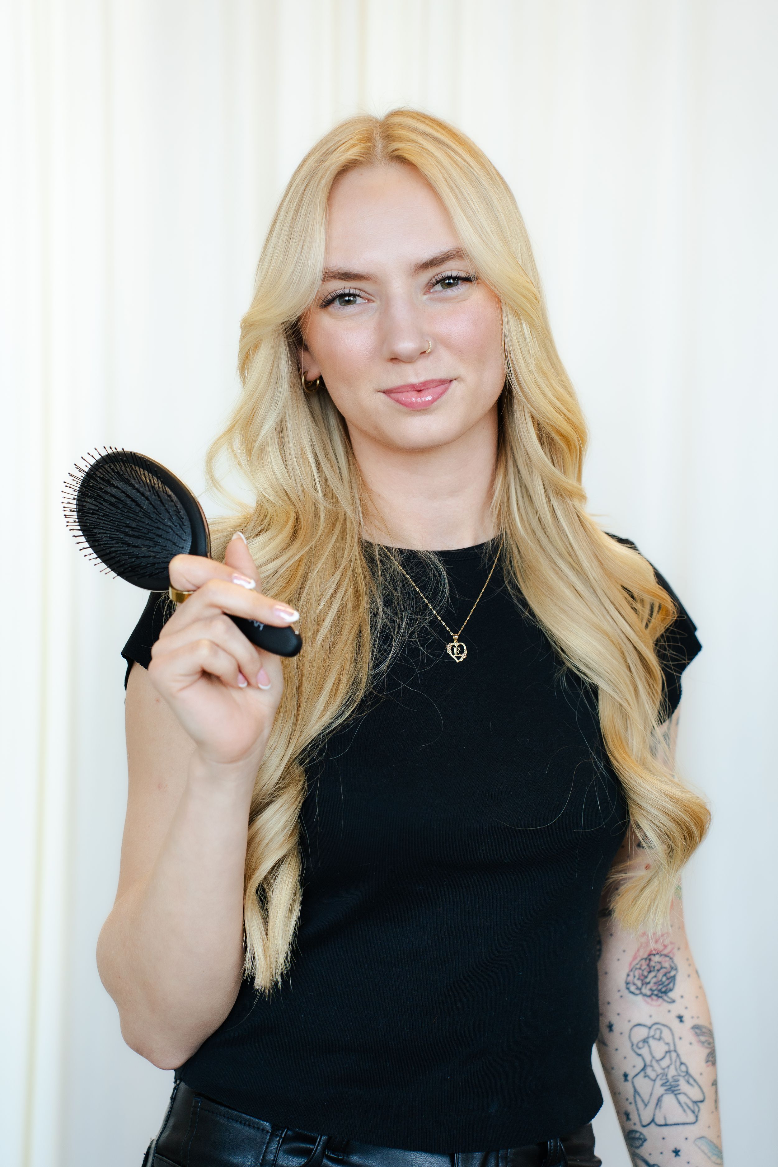 Blonde woman in black top holds hairbrush while showing arm tattoo against white backdrop.