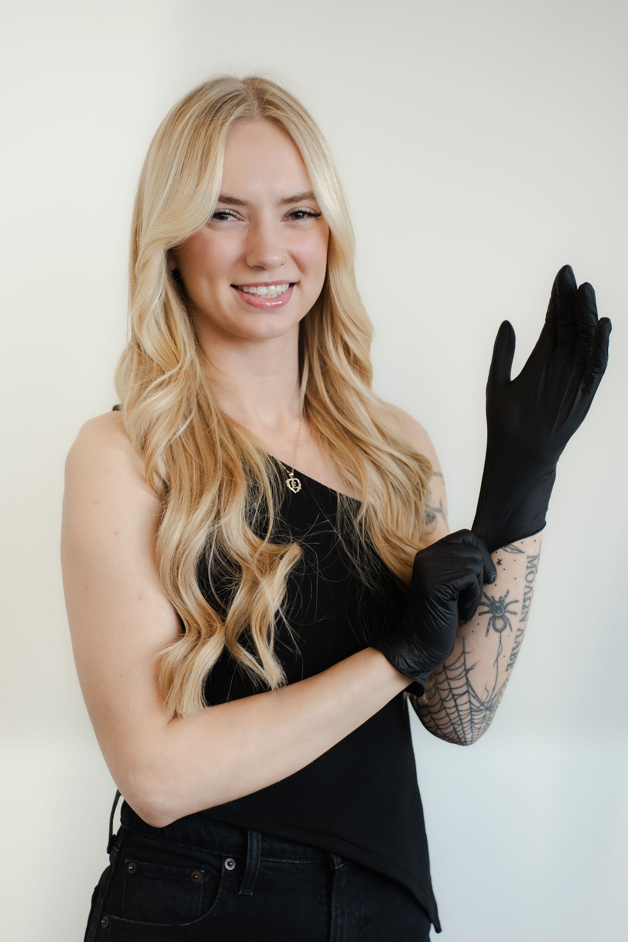 Professional portrait of a blonde woman in black attire putting on a black glove, with visible arm tattoo artwork.