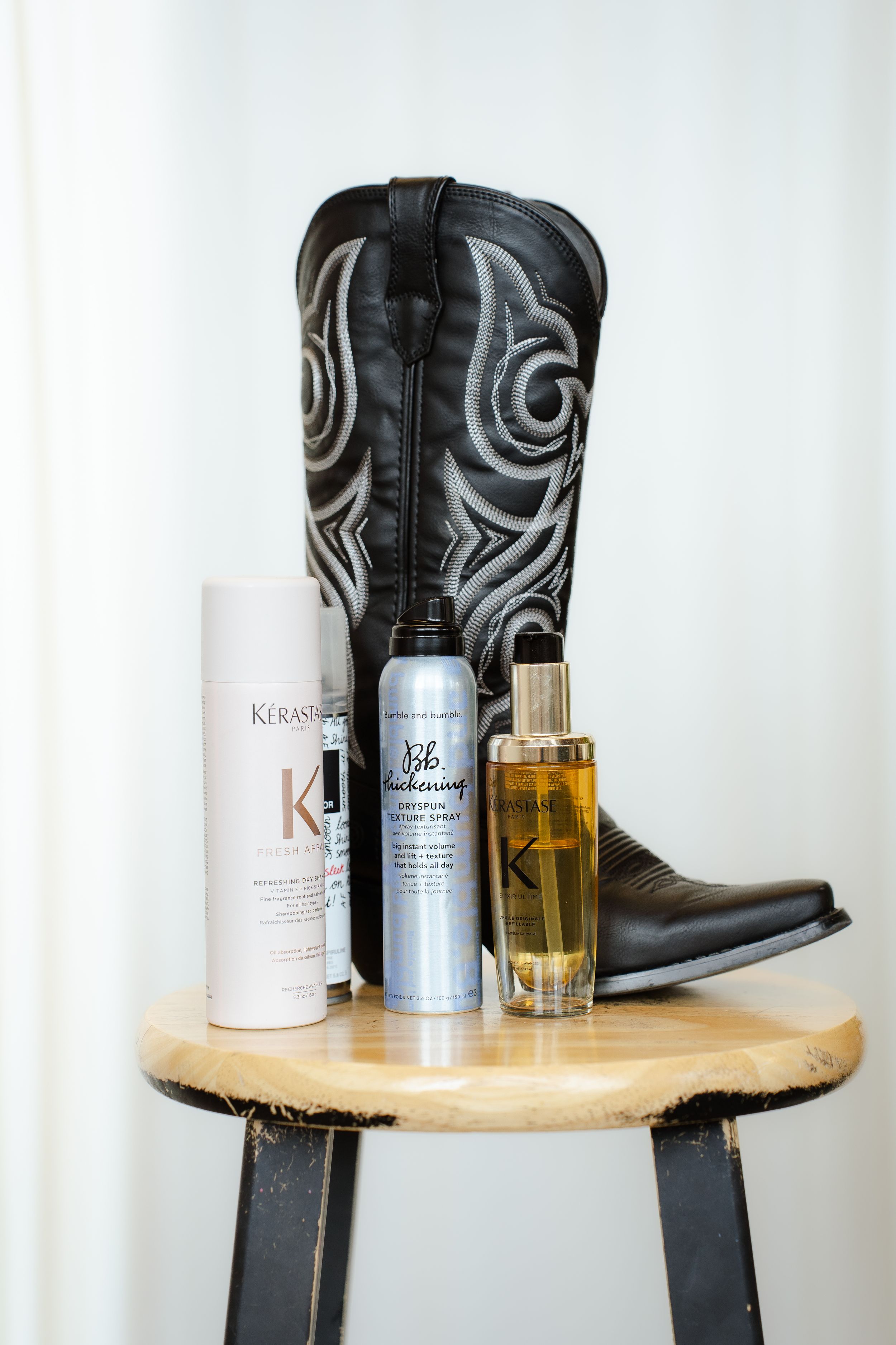 Black leather cowboy boots and beauty products arranged on a rustic wooden shelf against a white background.