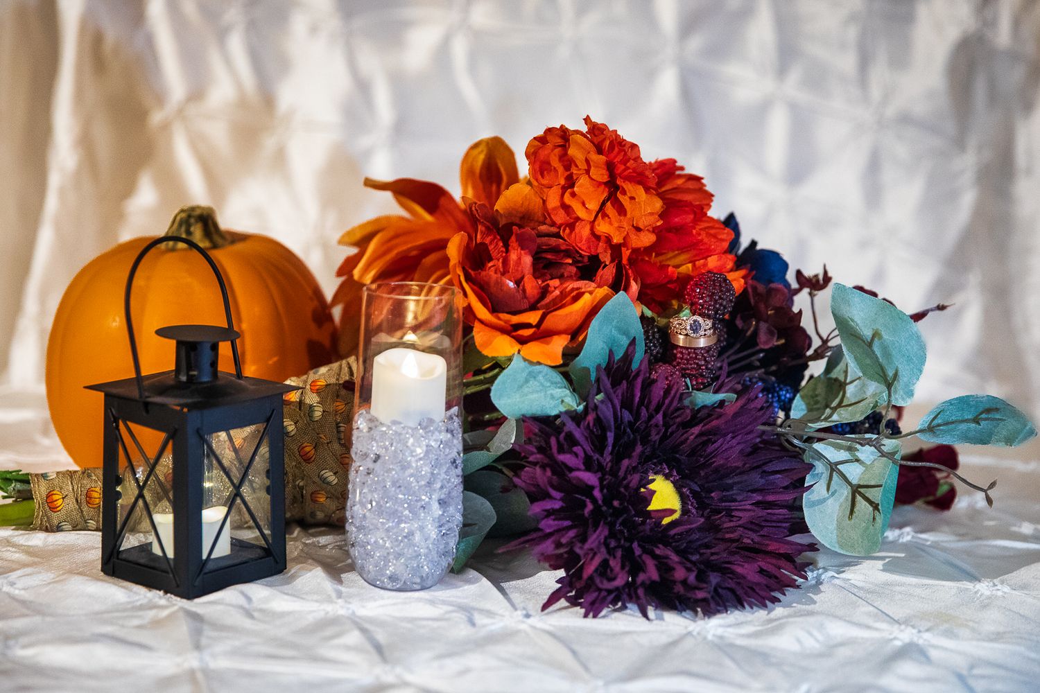 Halloween decorative display with orange pumpkins, purple flowers, lanterns and blue accents on white fabric background.