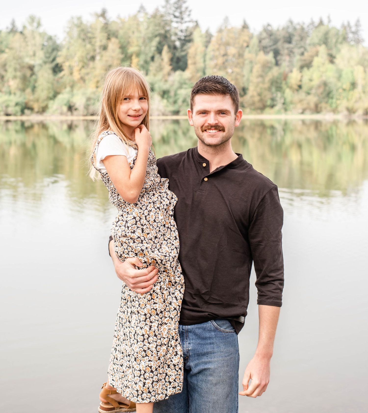 A couple poses together by a serene lake surrounded by evergreen trees on a bright sunny day.