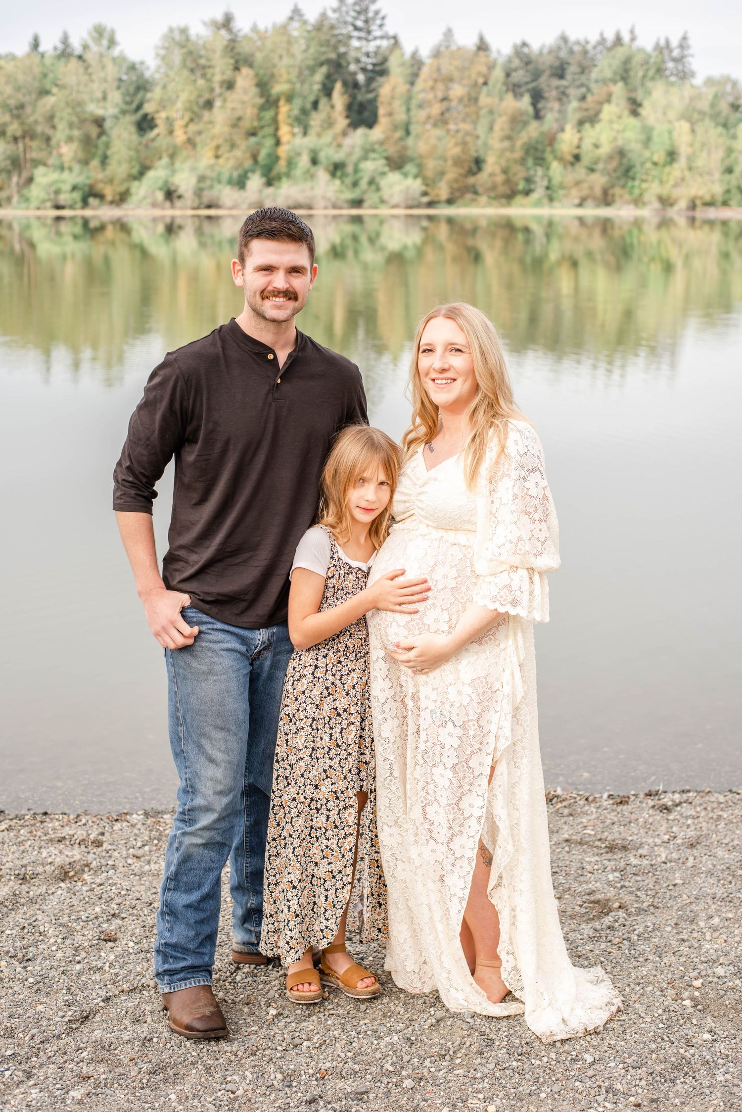 Family maternity photo session by a lakeside with people wearing coordinating brown and cream outfits.