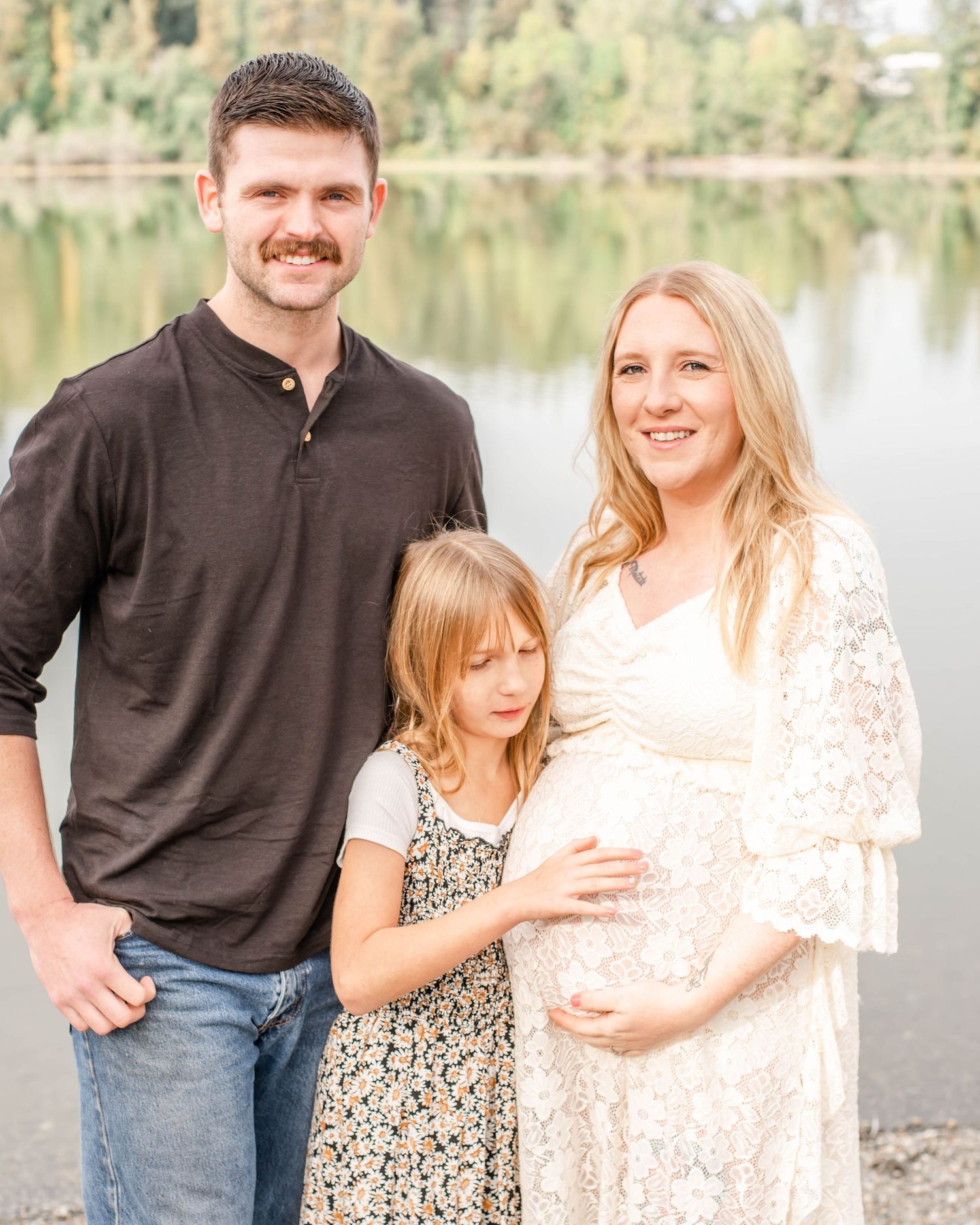 Family standing together near a serene lake surrounded by trees in warm evening light.