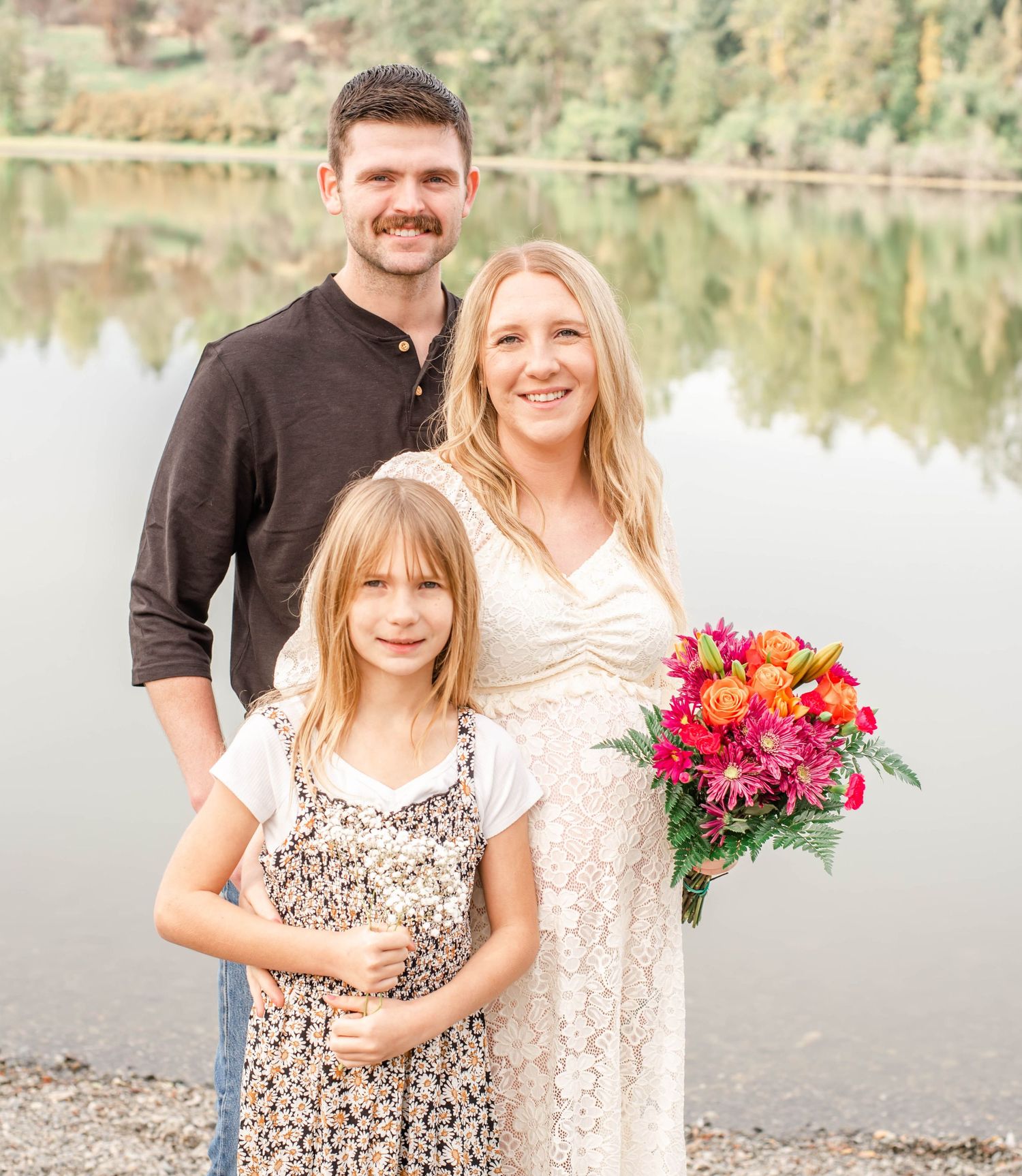 A family poses together by a serene lake with colorful floral bouquets and casual attire.