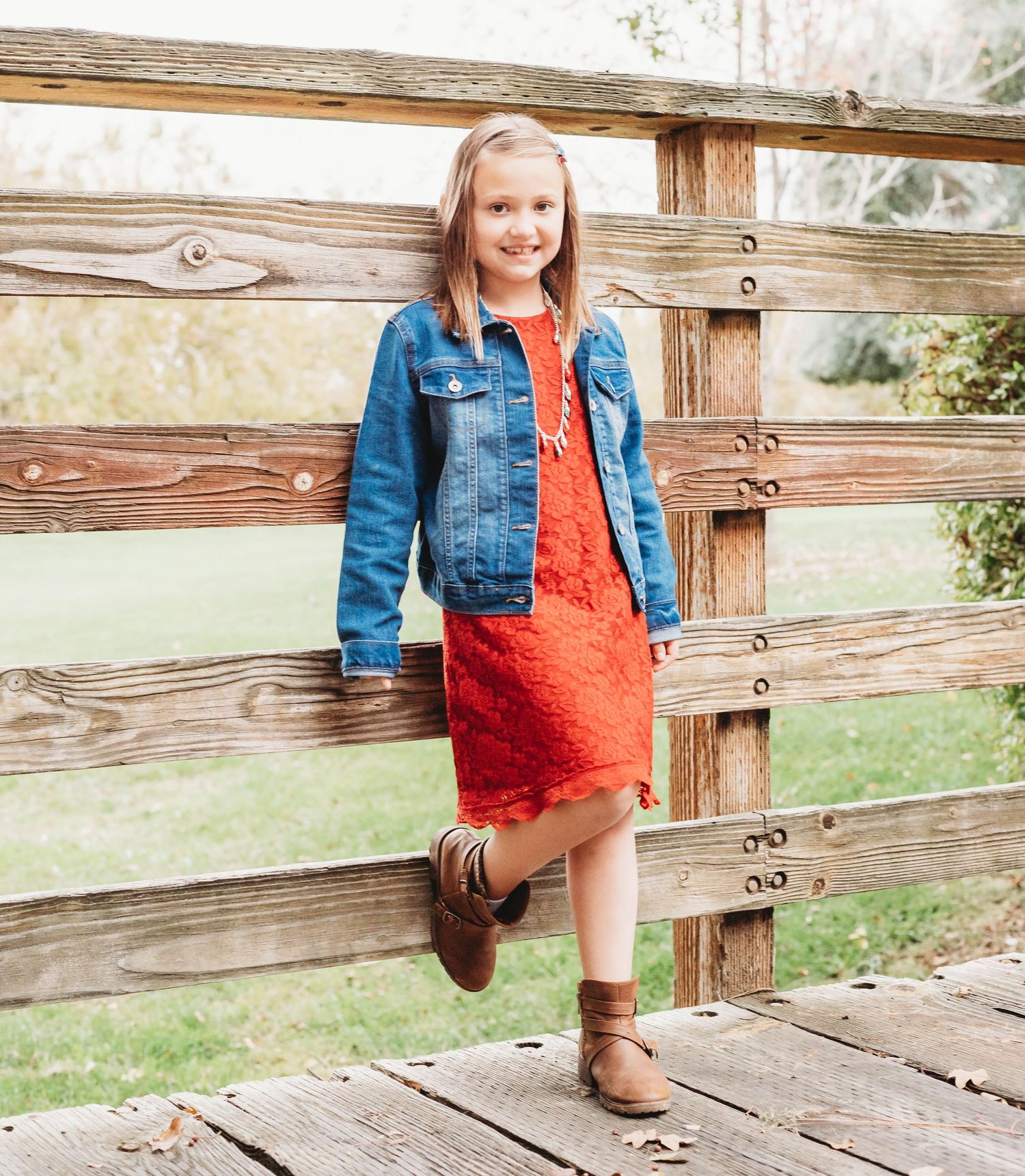 A person in a denim jacket and red dress sits on a rustic wooden deck railing in a countryside setting.