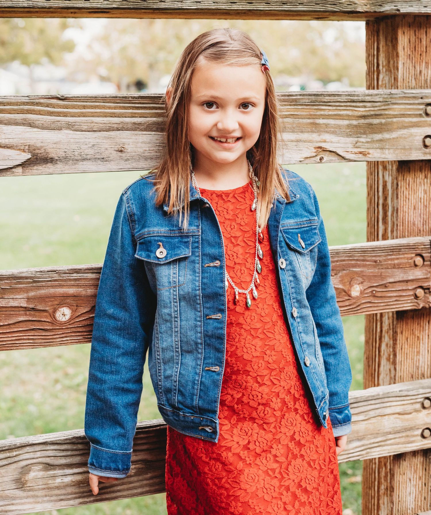 A young person in a red lace dress and denim jacket poses by a rustic wooden fence in an outdoor setting.
