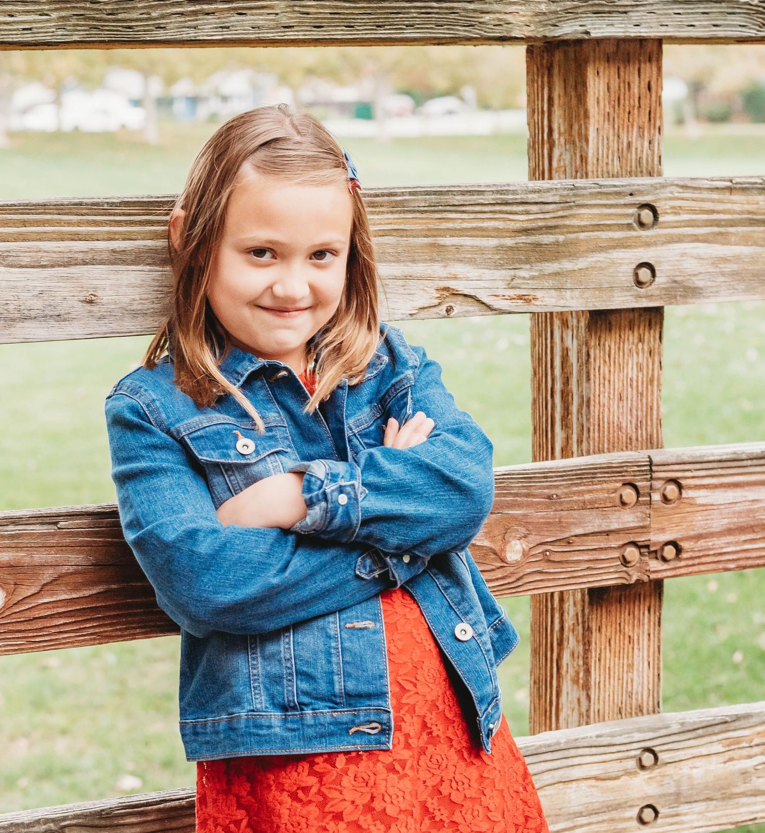 Young person in denim jacket and red dress poses against rustic wooden fence in outdoor setting.