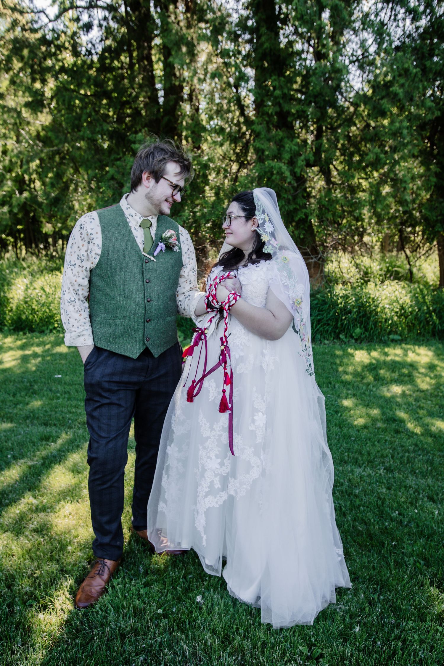A couple shares an intimate moment in a garden setting wearing wedding attire with a lace dress and green vest.