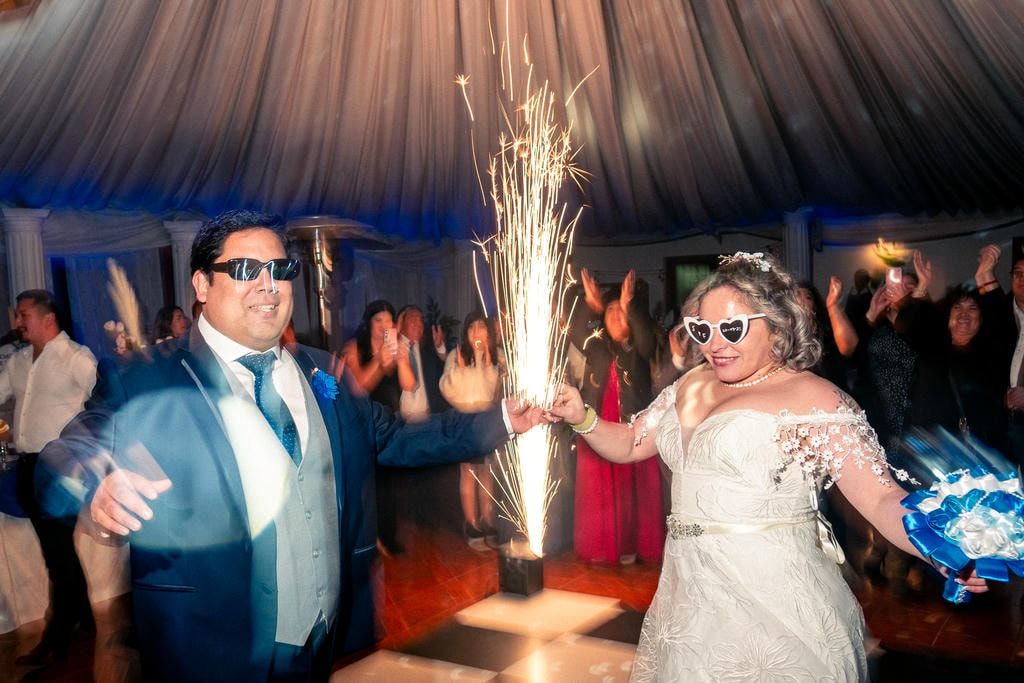 Recién casados bailando bajo bengalas en la recepción, con invitados celebrando en carpa.