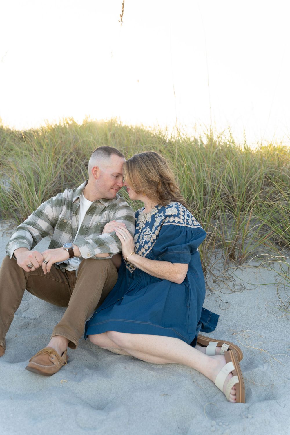 Romantic couple lying together on sandy beach at sunset wearing blue dress and plaid shirt.