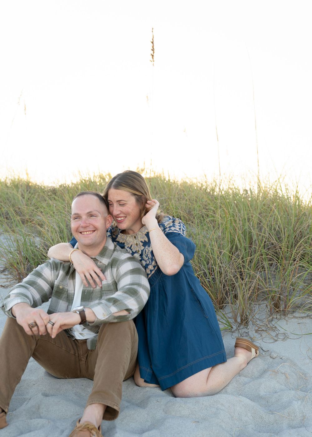 A couple embraces while sitting on sand dunes during a romantic beach photoshoot at golden hour.
