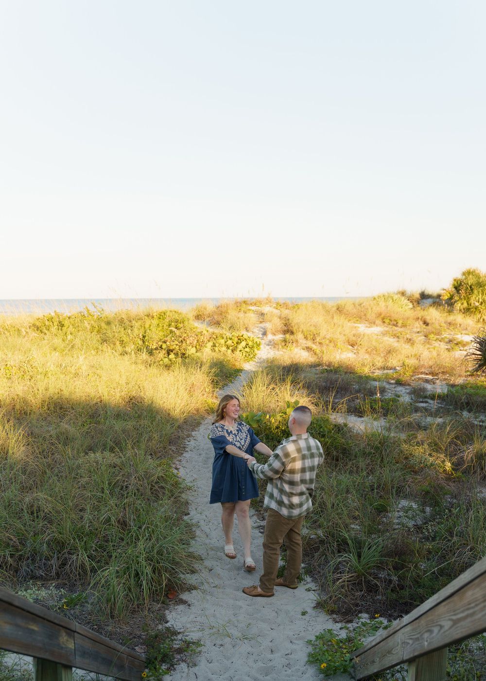 Two people stand together on a sandy beach path surrounded by dunes and coastal vegetation at sunset.