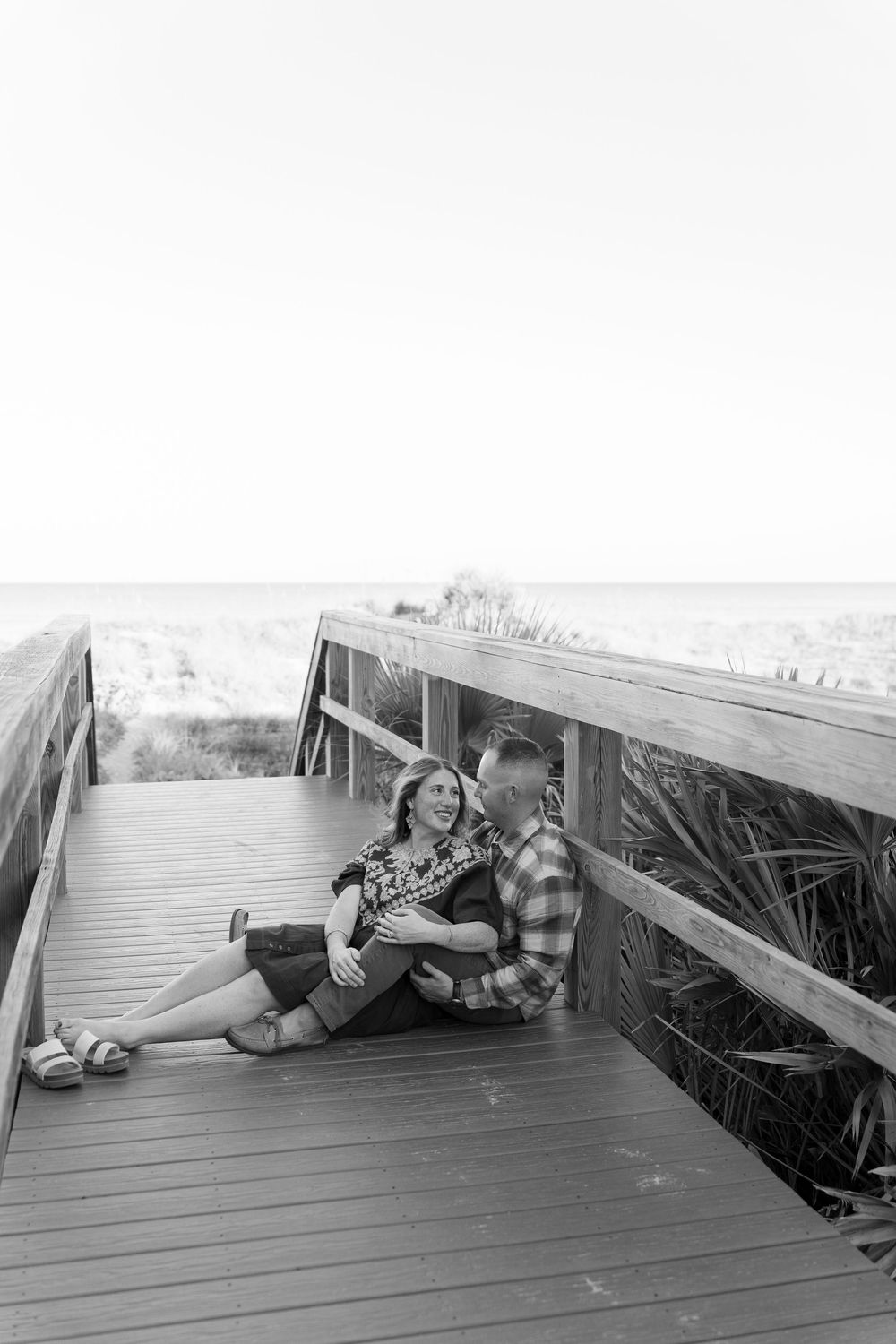 Couple sharing playful moments while lying on wooden boardwalk in black and white photography.