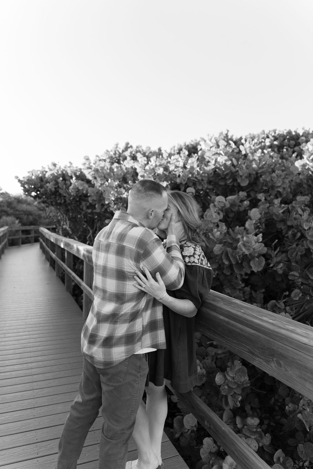 Couple embracing on wooden boardwalk surrounded by coastal vegetation.