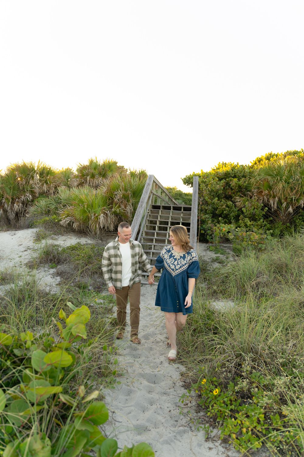 A couple walks together on a sandy beach path surrounded by coastal vegetation at sunset.