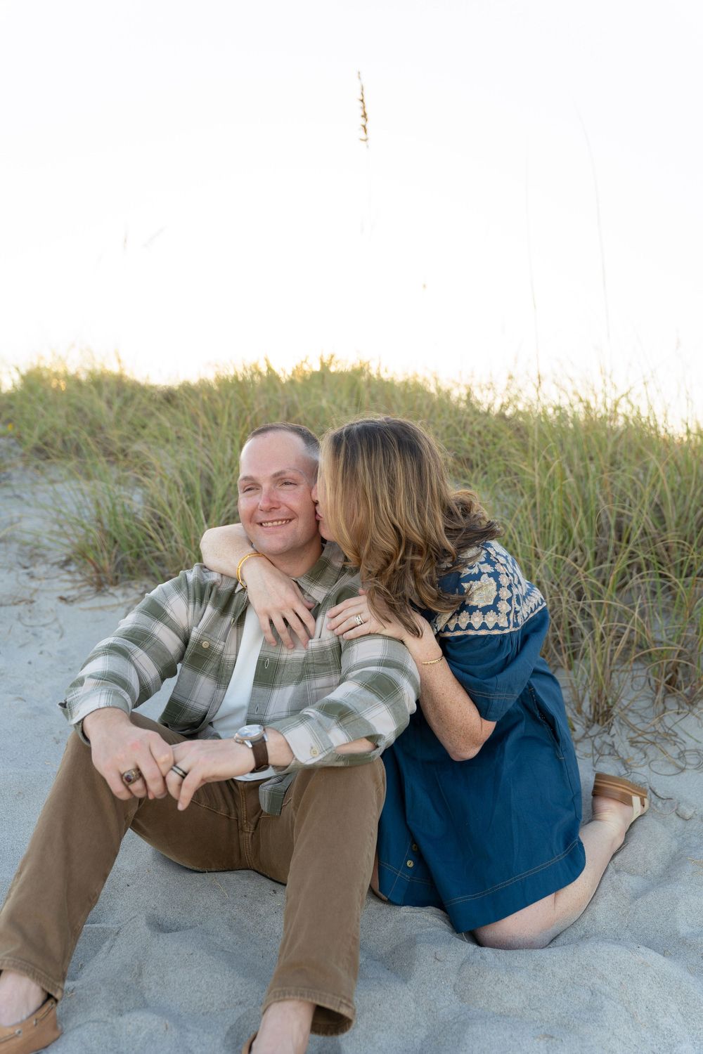 A couple shares an intimate moment sitting together on sandy dunes with beach grass in the background at sunset.