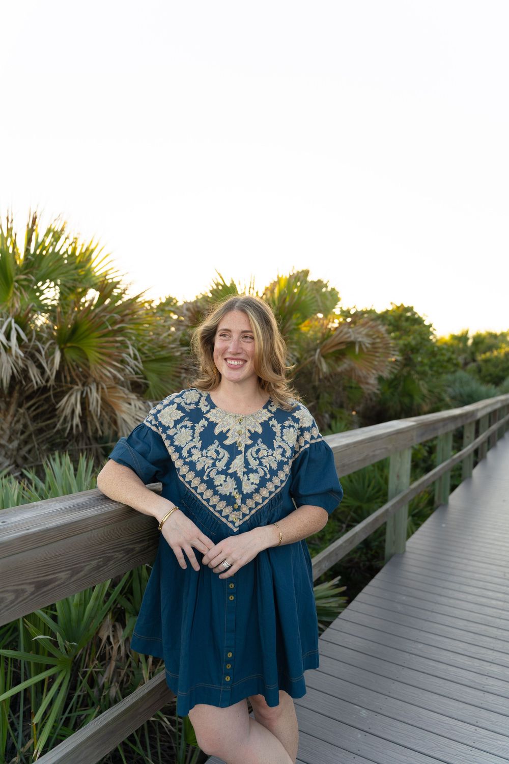 Woman in blue embroidered dress poses on wooden boardwalk surrounded by palm trees at sunset.