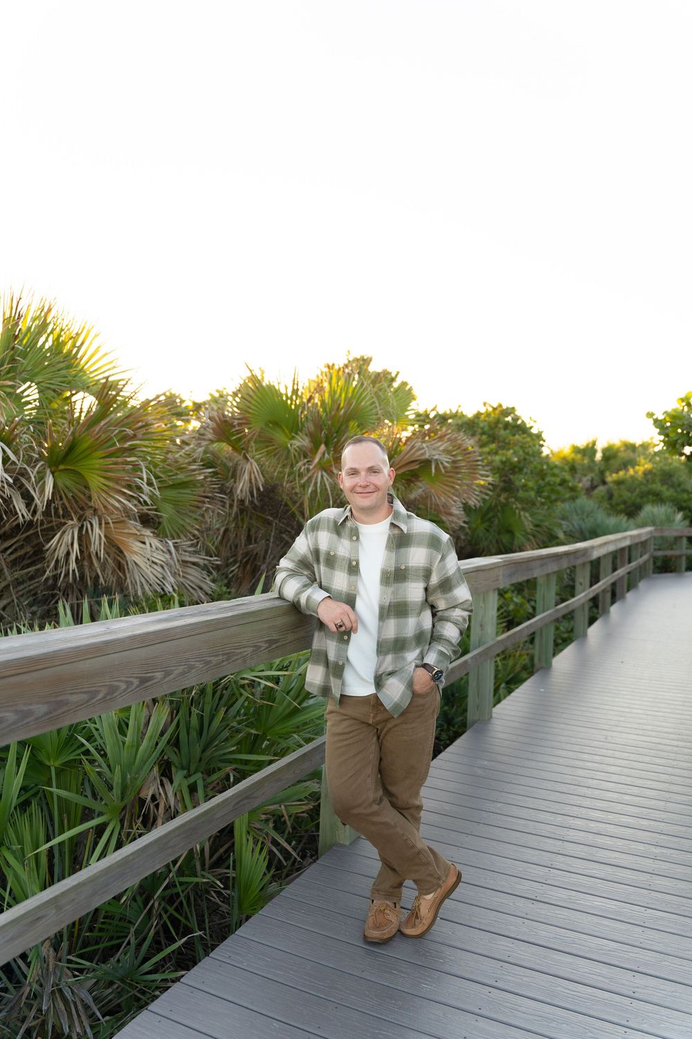 Person in plaid shirt and khakis poses on wooden boardwalk surrounded by tropical plants at sunset.