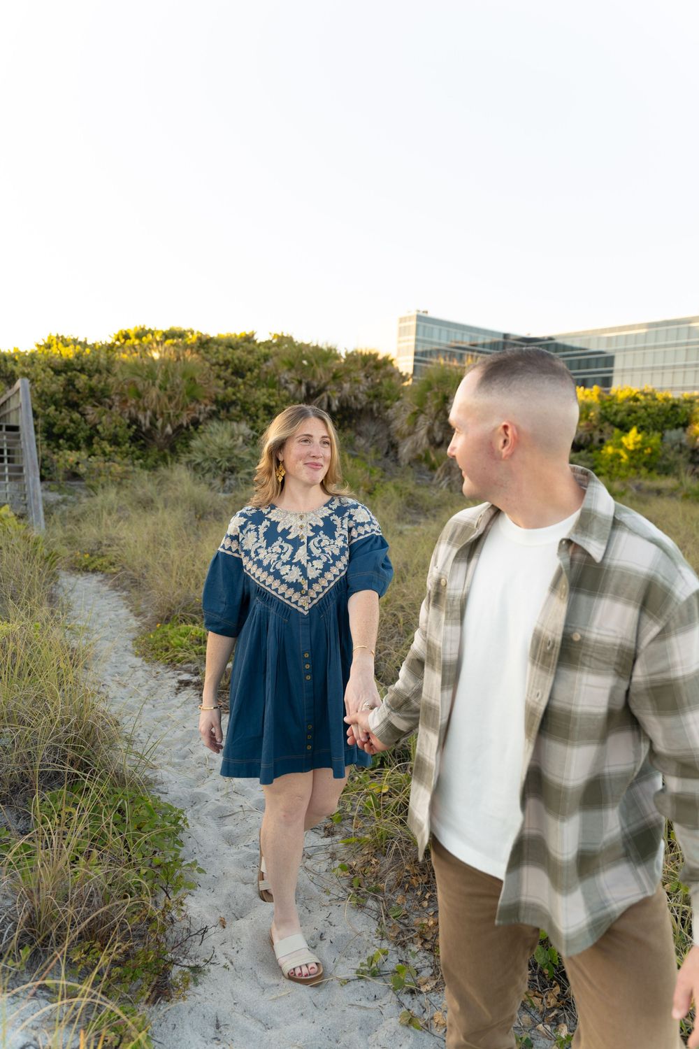 Couple walking together on sandy beach path at golden hour wearing blue dress and plaid shirt.