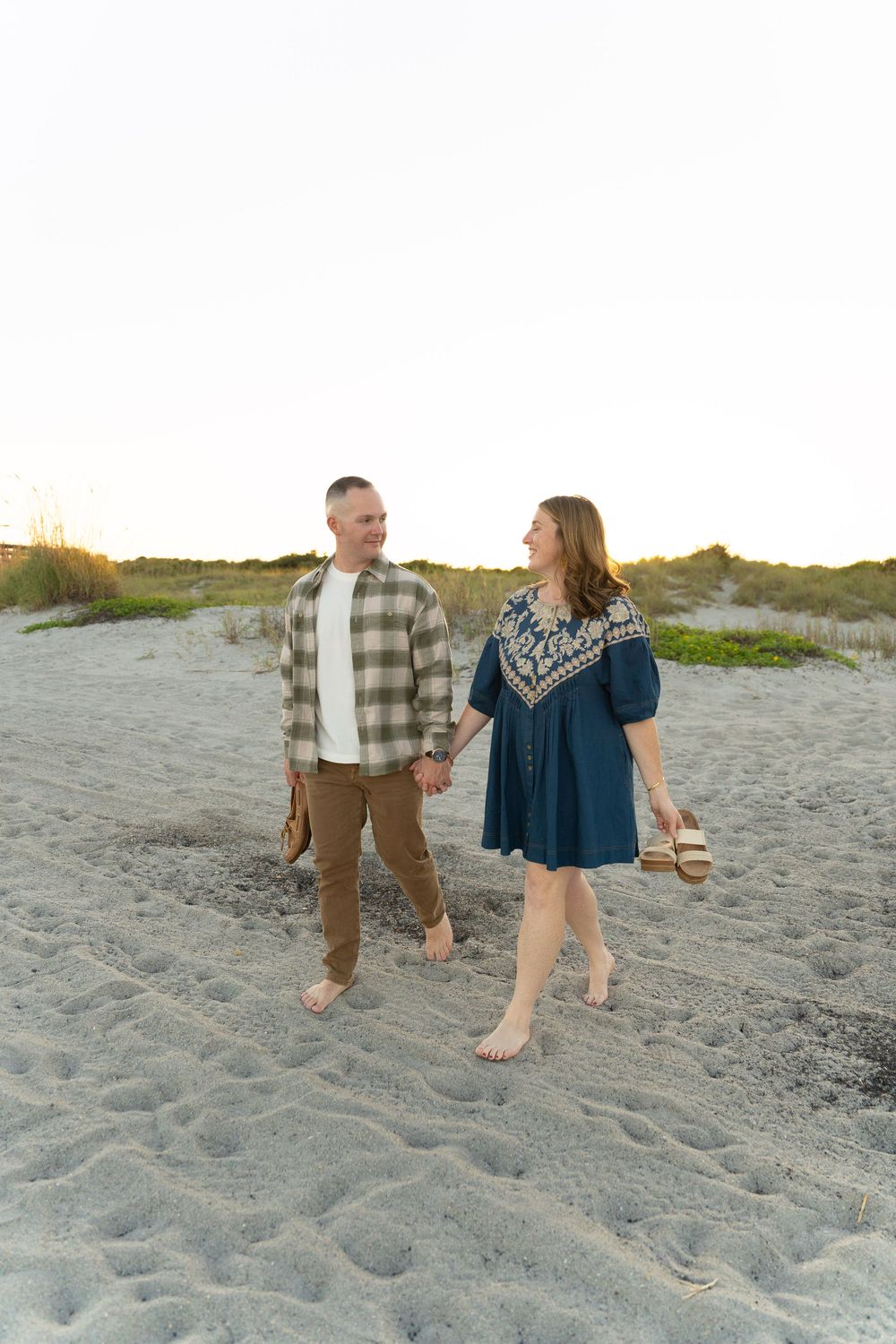 A couple walks hand in hand along a sandy beach at sunset wearing casual clothing and enjoying a romantic moment.
