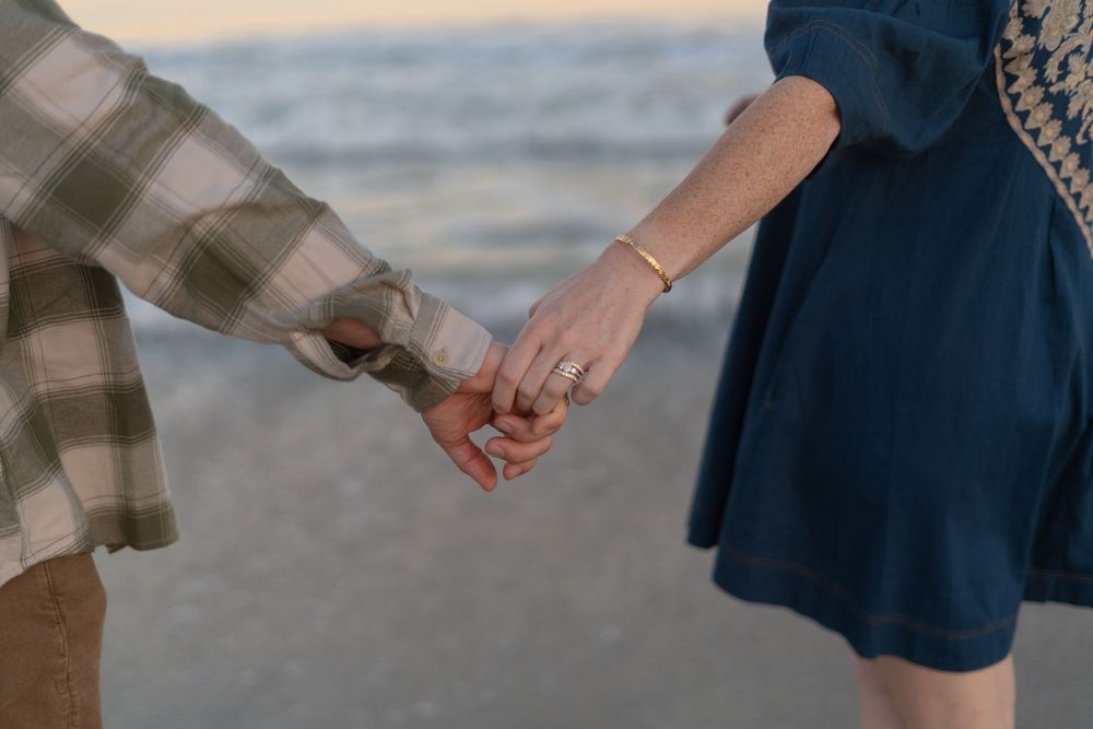 Two people holding hands on a beach wearing plaid and navy blue clothing against an ocean backdrop.