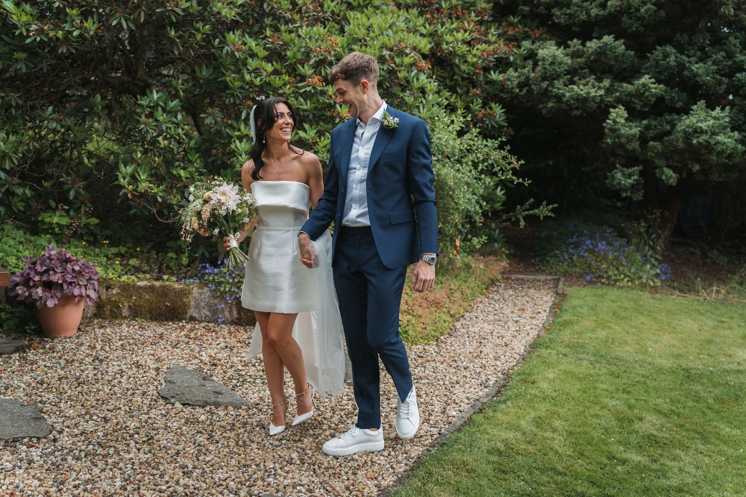 A couple walks together on a gravel path surrounded by lush greenery during an outdoor wedding celebration.