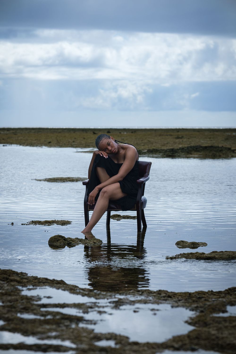 A person sits in a black chair placed in shallow water during low tide, with moody cloudy skies overhead.