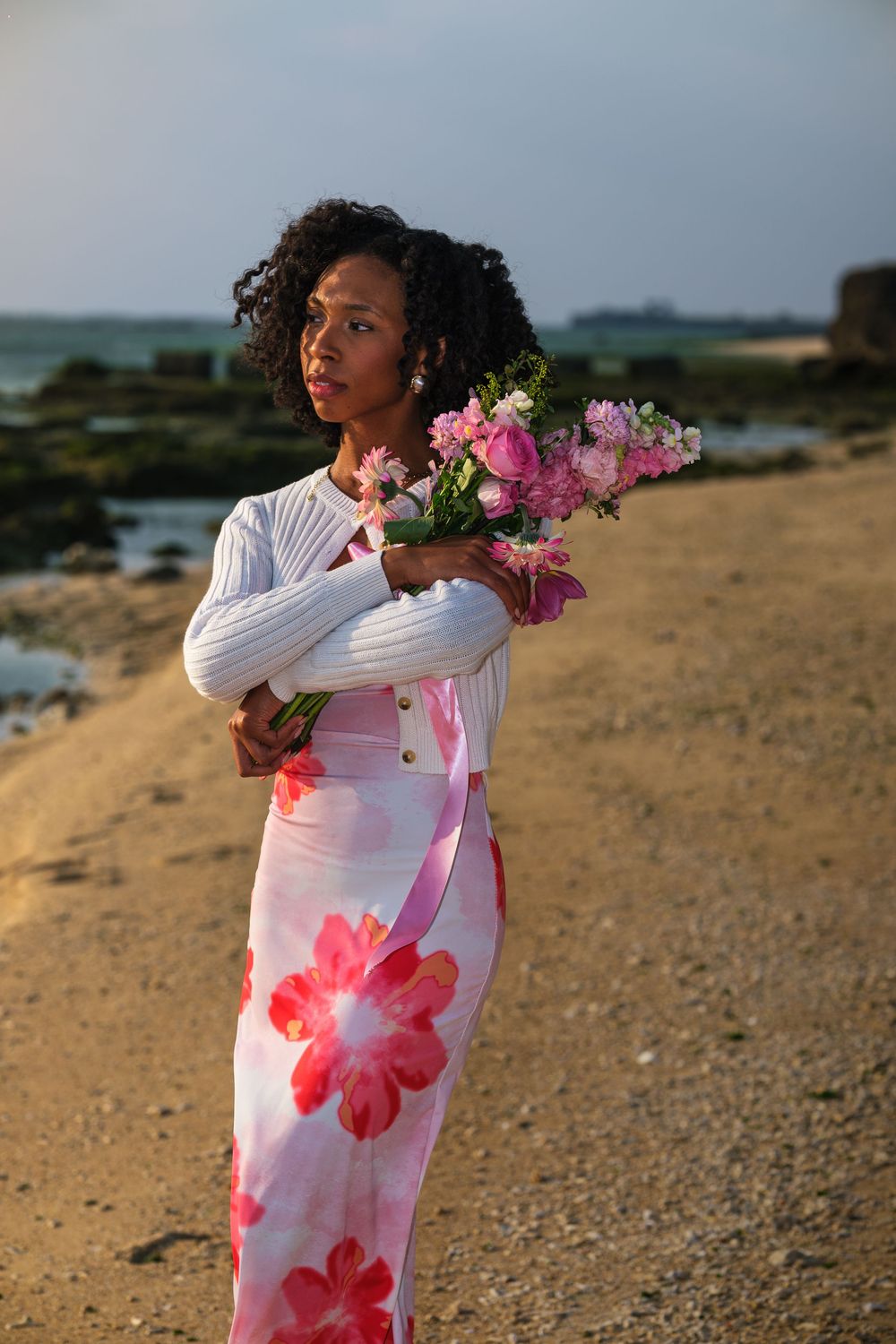 A person in a floral dress holds flowers while standing on a sandy beach at sunset.