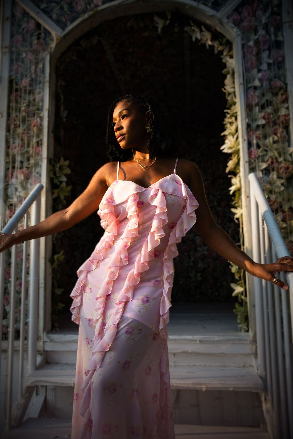 Elegant pink ruffled evening gown with spaghetti straps photographed in romantic garden archway during golden hour.