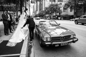 A classic Mercedes-Benz car parked on a city street with people standing nearby during a wedding celebration.