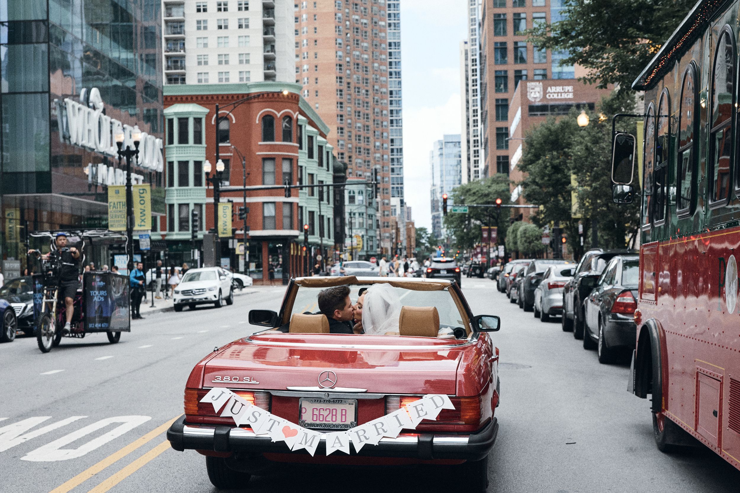 A vintage convertible decorated with wedding ribbons drives down a busy city street lined with tall buildings.
